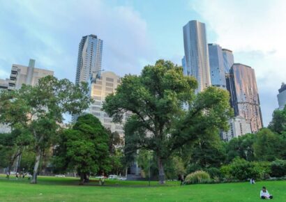 People relaxing on an open green lawn in a Melbourne park with mature trees and CBD high-rises rising in the background under a partly cloudy sky.