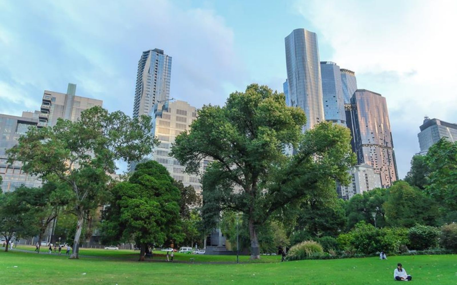 People relaxing on an open green lawn in a Melbourne park with mature trees and CBD high-rises rising in the background under a partly cloudy sky.