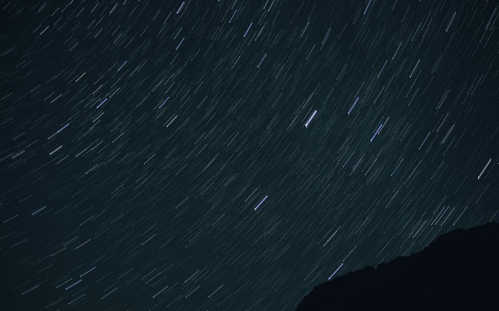 Alt text: Curved star trails sweep across a dark teal sky in a long-exposure photograph with a mountain ridge silhouetted in the lower right corner