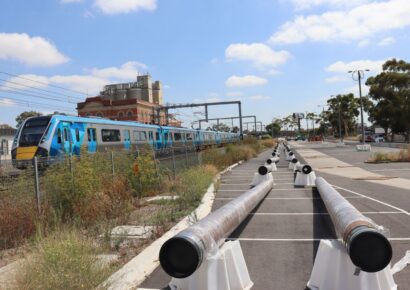 Steel pipeline sections laid out in Albion Station car park as part of early works for Melbourne Airport Rail Stage 1, with a Metro train passing on the adjacent rail corridor