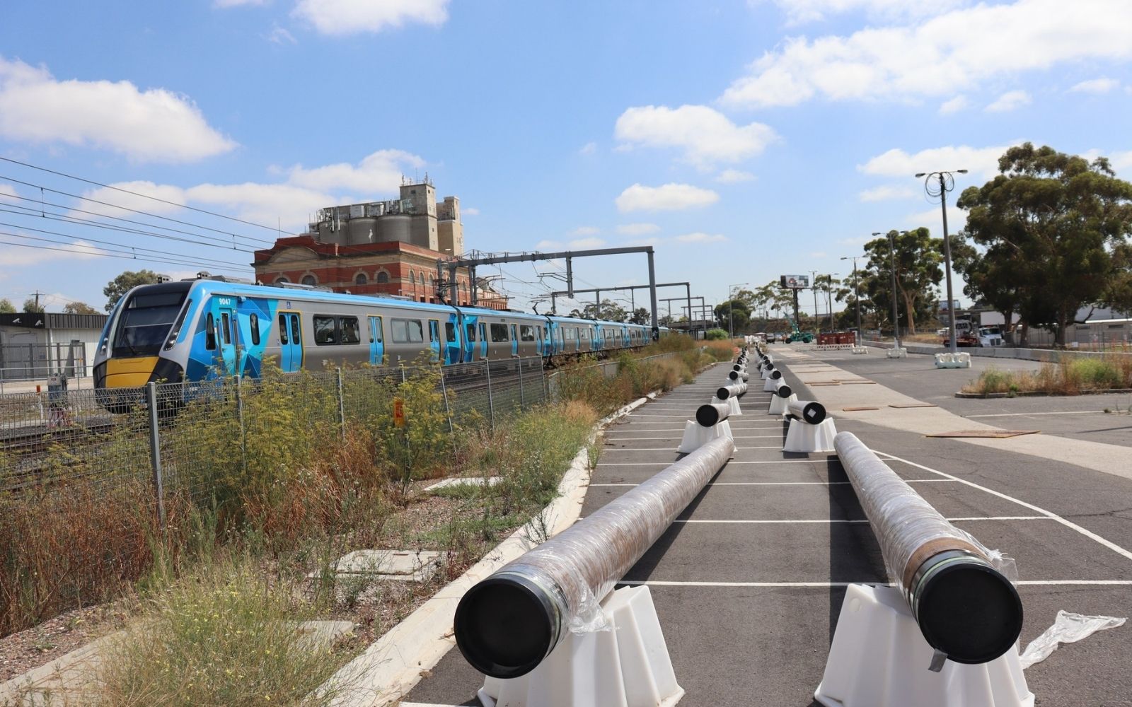 Steel pipeline sections laid out in Albion Station car park as part of early works for Melbourne Airport Rail Stage 1, with a Metro train passing on the adjacent rail corridor