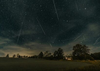 Alt text: Meteor streaks and satellite trails cross a star-filled night sky above a rural landscape with silhouetted trees and a warmly lit house