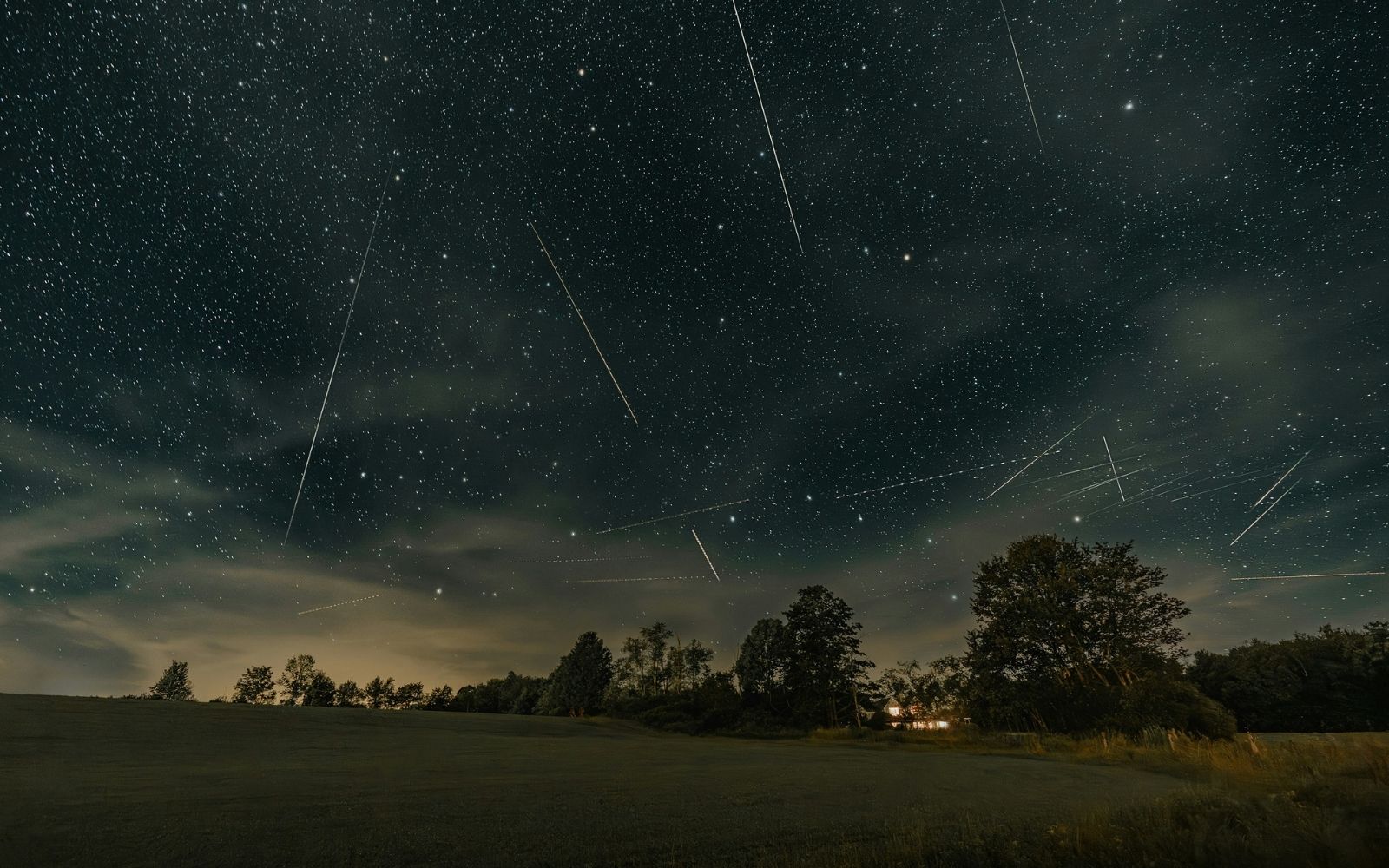 Alt text: Meteor streaks and satellite trails cross a star-filled night sky above a rural landscape with silhouetted trees and a warmly lit house