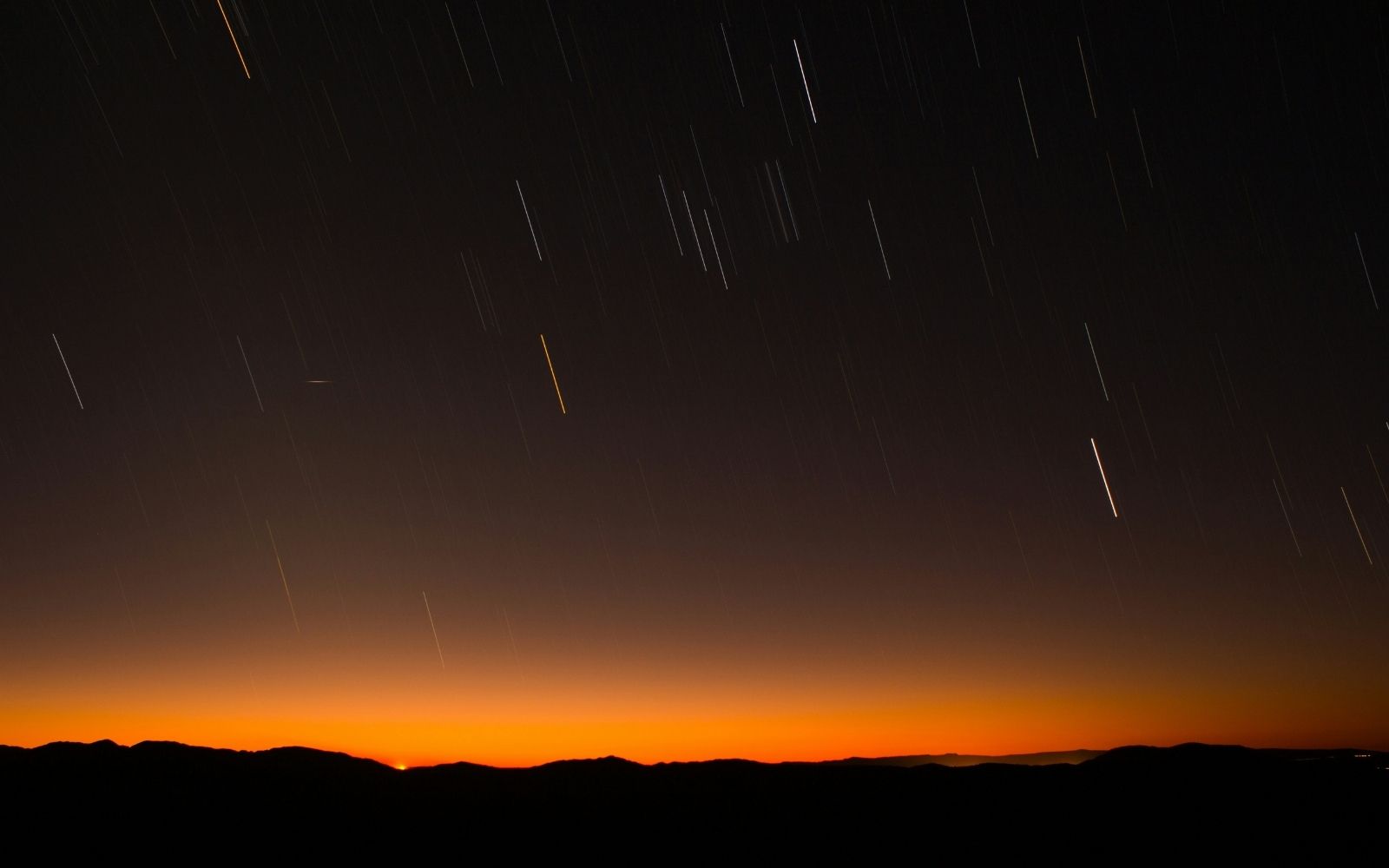 Alt text: Long-exposure star trails fall like rain across a dark sky above a mountain range silhouetted against a deep orange sunset glow