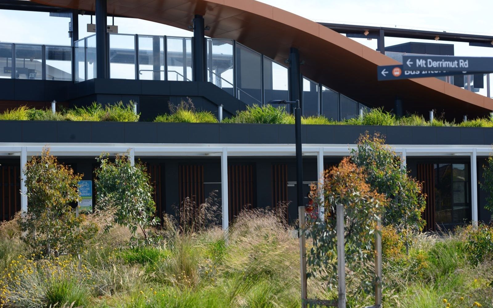 Alt text: Close view of Deer Park Melbourne train station showing native plants spilling from the rooftop garden above the station building, with wayfinding signage pointing to Mt Derrimut Road and the bus interchange