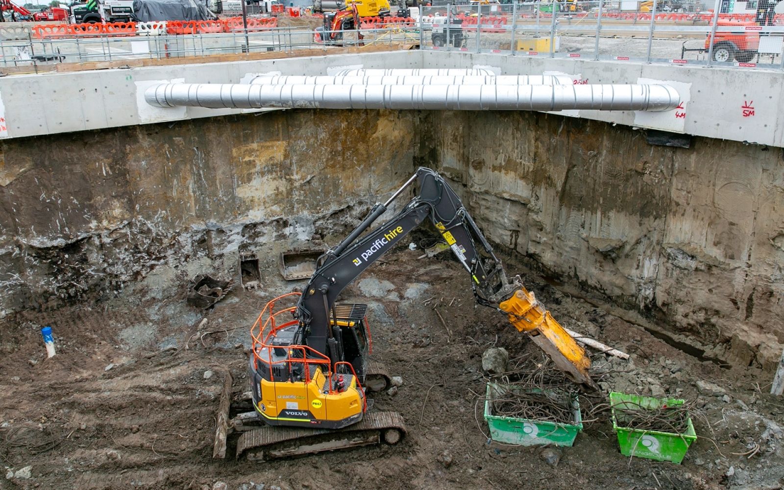 An excavator operates at the bottom of the deep launch shaft at the Suburban Rail Loop's Clarinda construction site, with reinforced concrete walls and steel bracing visible above.