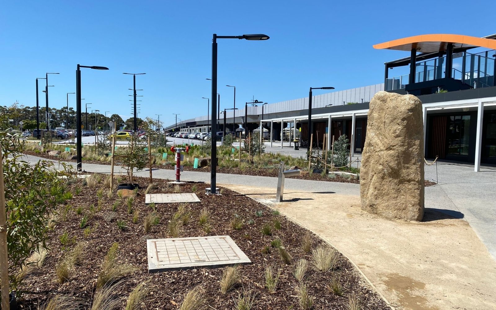 Alt text: The landscaped forecourt at Deer Park Melbourne train station, showing native plantings, young trees and one of three giant boulders oriented toward nearby mountain ranges