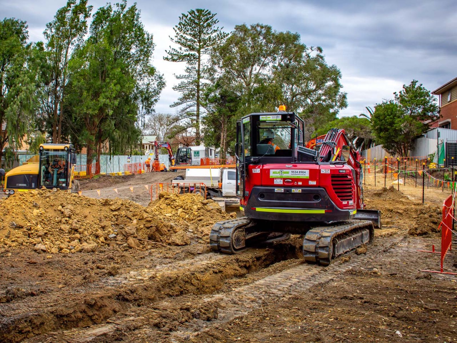An excavator clears ground at a Suburban Rail Loop East construction site in suburban Melbourne, with earthworks underway beside local homes and trees.