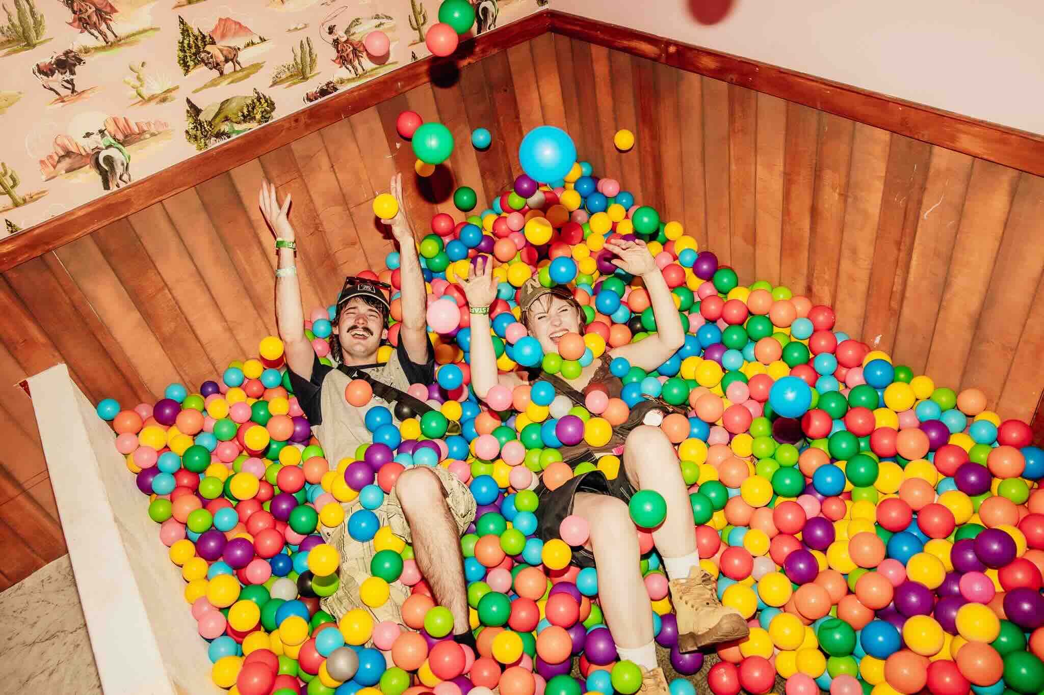 Two festival-goers laugh in a colourful ball pit under cowboy-print wallpaper at Beyond The Valley, part of the site around the BTV lineup.